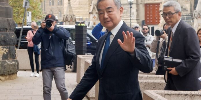 Man in a dark pinstripe suit and blue tie waves at photographers as cameras flash outside a historic building onlookers present.