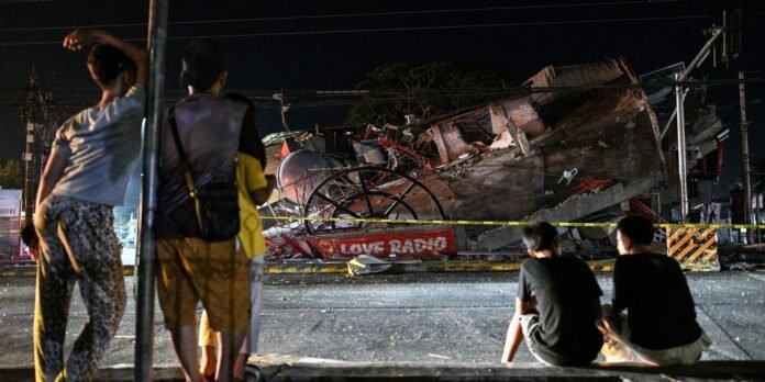 Nighttime street view of a collapsed building behind yellow caution tape, with a red 'LOVE RADIO' banner and onlookers in the foreground.