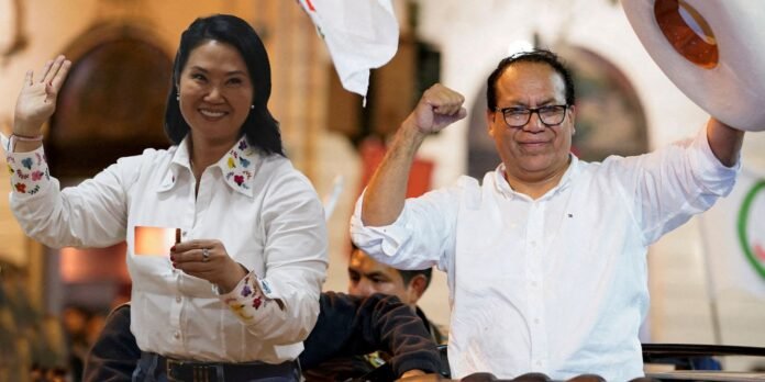 Two people celebrate: a smiling woman in a white embroidered shirt waves and holds a small card, as a man beside her pumps his fist and lifts a hat in triumph.