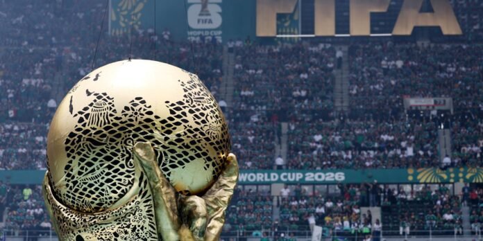 FIFA World Cup trophy close-up in gold with intricate lattice pattern, held by a gloved hand as a packed stadium watches in the background.