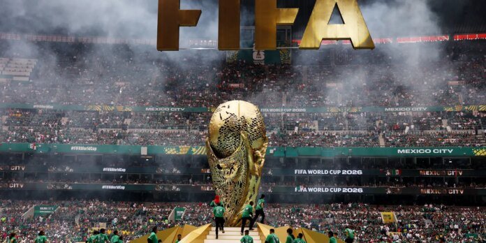 Big gold FIFA letters hang above a crowded stadium during the FIFA World Cup 2026 with a large decorative trophy centerpiece on a stepped platform, fans in green filling the stands, and smoke in the air.