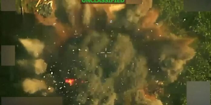 Aerial view through a targeting scope with a central crosshair, showing smoke plumes and small explosions on the ground; UNCLASSIFIED label at the top.