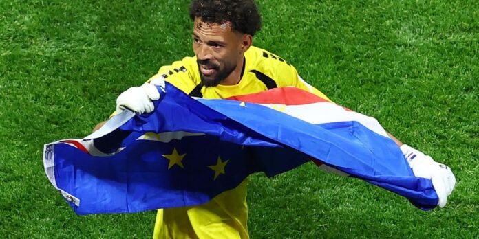 Goalkeeper in a yellow kit waving the Cape Verde flag on a green pitch, celebrating victory.