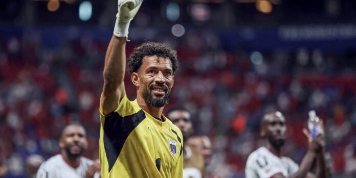 Soccer goalkeeper in a yellow and black kit raises a gloved hand in celebration on the pitch, stadium crowd blurred behind him.