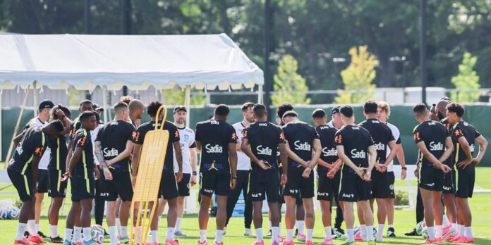 Soccer players in black training kits form a circle around a coach on a sunny practice field, with a yellow training mannequin nearby under a white tent.
