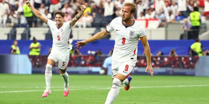 Two England soccer players in white jerseys celebrate a goal on a green field, with a cheering crowd in the stands behind them (one wearing armband and number 9).