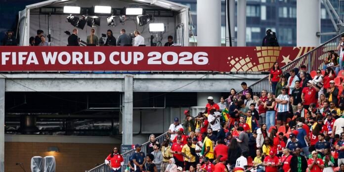 Banner reading 'FIFA World Cup 2026' over a stadium with crew and lights on a platform above a cheering crowd.