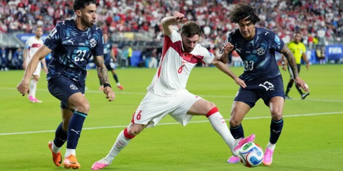 Soccer action: a white-uniformed player with red trim (number 6) challenges two defenders in navy kits for the ball on a green pitch with a crowded stadium backdrop.