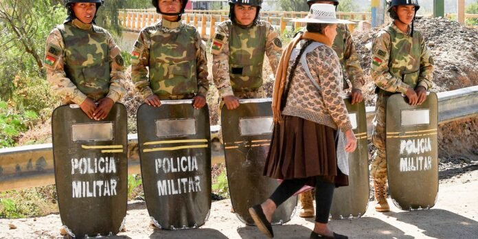 Camouflage-clad soldiers with riot shields reading 'Policia Militar' stand in a line behind a barrier as a woman in a knitted sweater passes by on a dirt road.