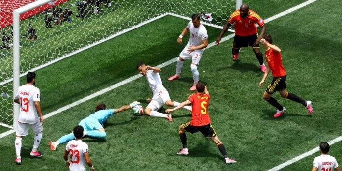 Soccer goalmouth scramble with white-team attackers and red-defenders converging as the ball sits near the goalkeeper in blue.