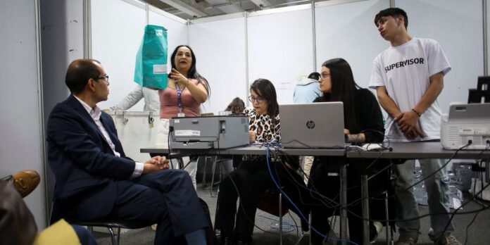 A woman stands and gestures with a turquoise bag while seated colleagues work on laptops at a booth table in a busy office-like setting.