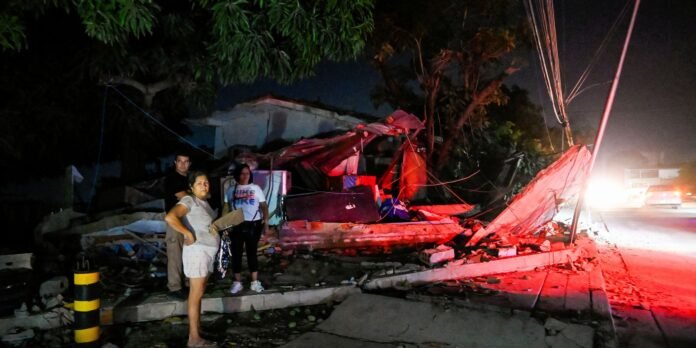 Nighttime scene of a collapsed building with rubble along the street; a small group of bystanders stands near the debris as red emergency lights coat the area in red.