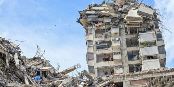 A man stands amid rubble of a destroyed multi-story building with exposed rebar against a blue sky.