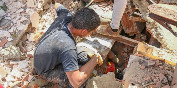 Workers in dusty rubble carefully breaking through debris to reach a collapsed area; one wears a red helmet below, others assist from above.