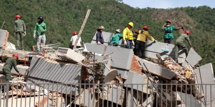 Rescue workers in hard hats and safety gear atop a collapsed building, clearing debris against a hillside backdrop.