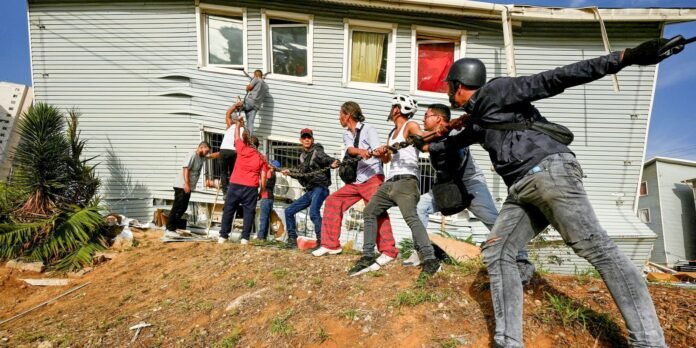 People in a disaster drill pull on a rope attached to a man climbing a damaged house, simulating a rescue scenario on a hillside.
