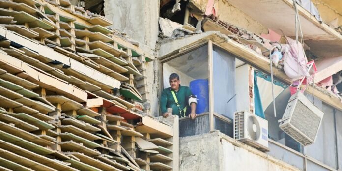 Man in a green shirt stands on a damaged balcony, surrounded by concrete rubble and stacked boards.
