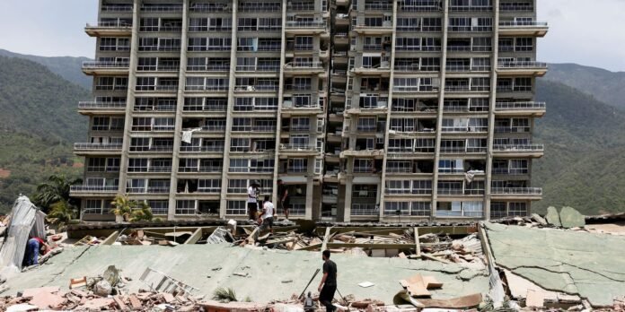 Collapsed high-rise building with shattered facade; people forage among rubble in the foreground against a mountainous backdrop.