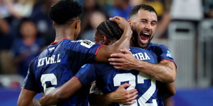 Three soccer players in blue jerseys embrace in celebration on the pitch after a goal.