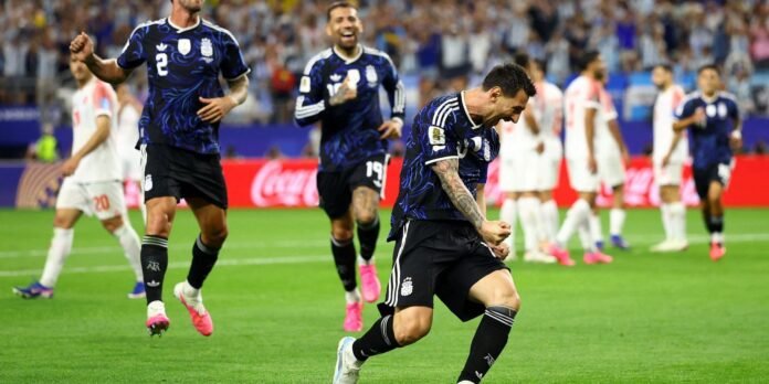 England football players in dark blue jerseys celebrate a goal on a green pitch, with one player in the foreground pumping his fist and others joyfully celebrating in the background.