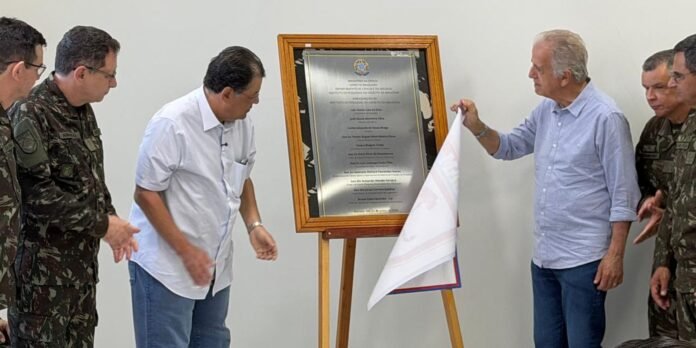 Group of men, including soldiers in camouflage, unveiling a metal commemorative plaque on a wooden easel during a ceremony.