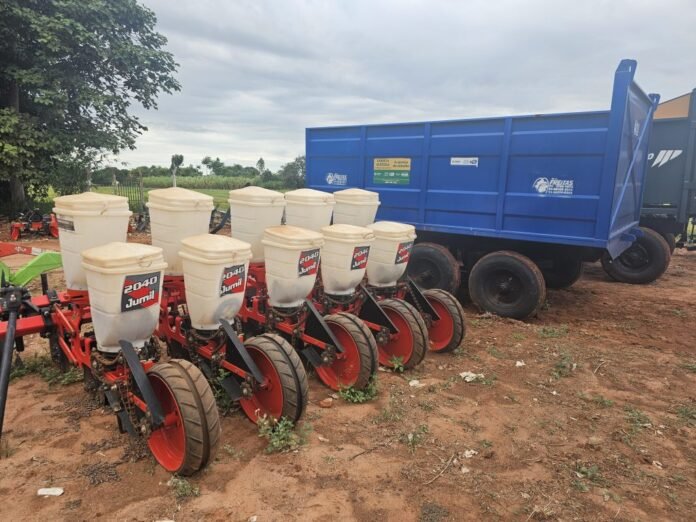 Row of red planter units with white seed hoppers (labeled 2040 Jumi) beside a blue grain trailer on a dirt field.