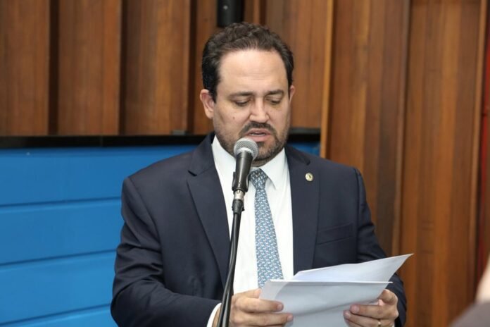 Man in a dark suit and patterned tie reads from papers into a microphone during a formal speech about to deliver remarks to an audience.