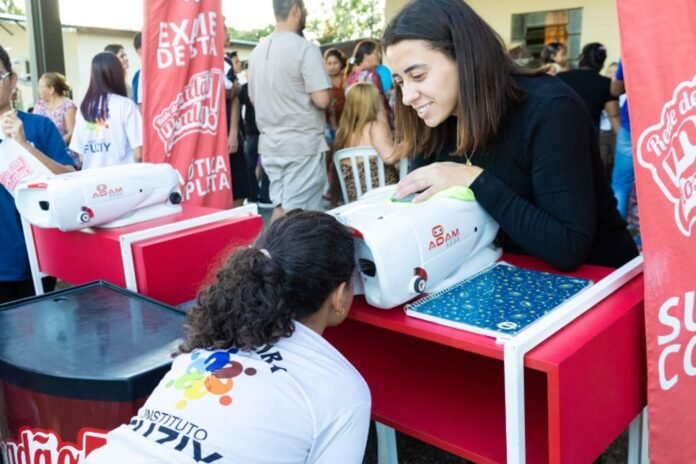 Smiling woman demonstrates a white device at a red booth to a young girl at an outdoor community event.
