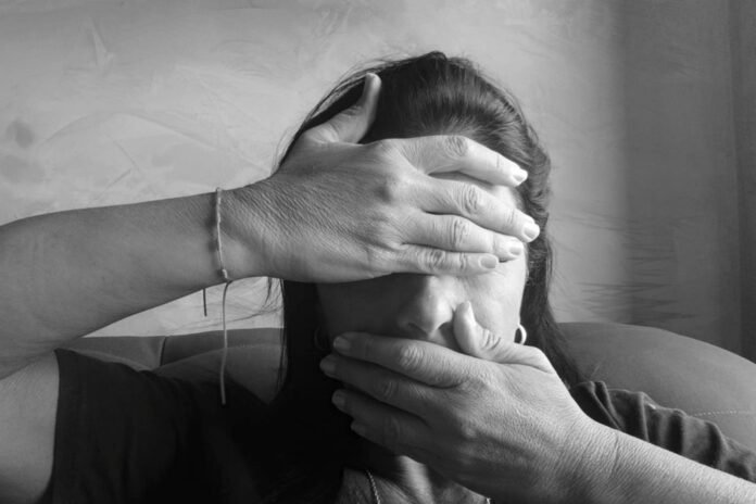 Woman covers her face with both hands, seated on a couch, conveying distress or overwhelm (black-and-white photo).