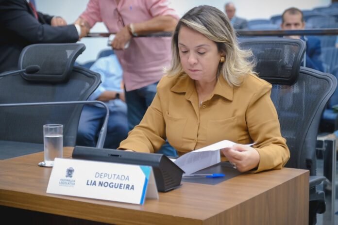 Woman in a tan blouse seated at a desk, reviewing papers in a legislative meeting room.