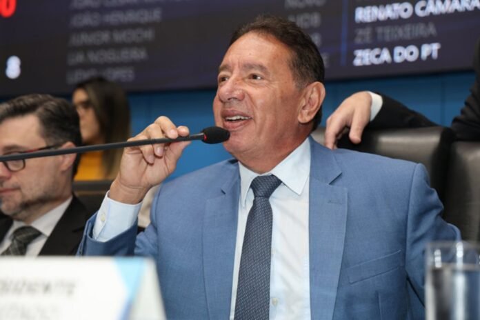 Man in a blue suit speaks into a microphone at a conference panel; colleague sits nearby in the background.