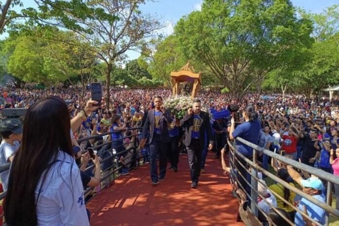 Casket carried on shoulders during outdoor funeral procession through a crowded park, with mourners and photographers nearby.