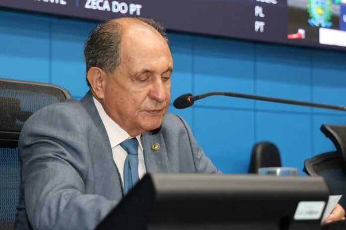 Older man in a gray suit and blue tie speaks into a microphone at a panel.