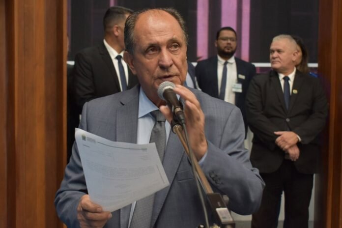 Man in a gray suit speaks into a microphone while holding a document, with other suited individuals in the background at a formal event.