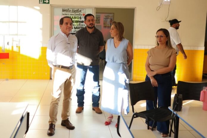 Four adults stand in a sunlit school hallway with yellow tile walls and a postered door in the background.
