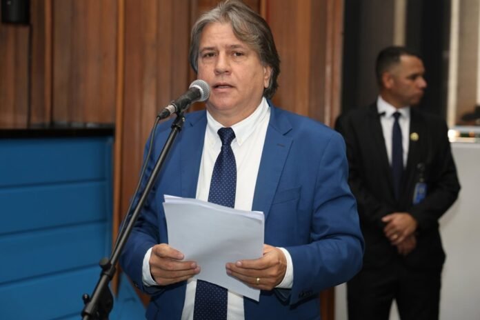 Man in a blue suit and polka-dot tie speaks into a microphone while holding papers at a formal event, with a security guard in the background.