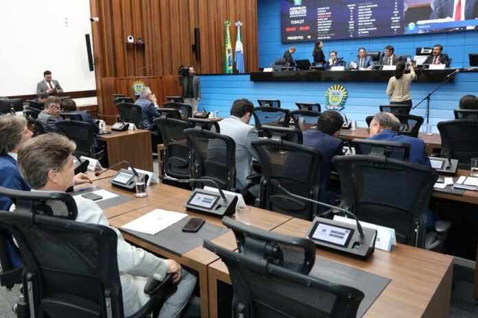 Council chamber scene with officials seated at curved desks facing a blue backdrop, a large screen displaying information, and flags behind a raised dais.