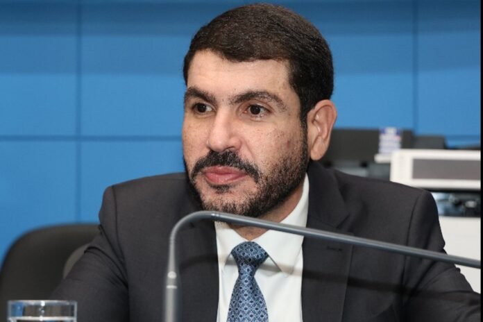 Man in a dark suit and blue tie speaks into a microphone at a formal meeting.