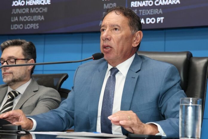 Man in a blue suit speaks into a microphone at a press conference, with a colleague beside him and a blue backdrop behind them.