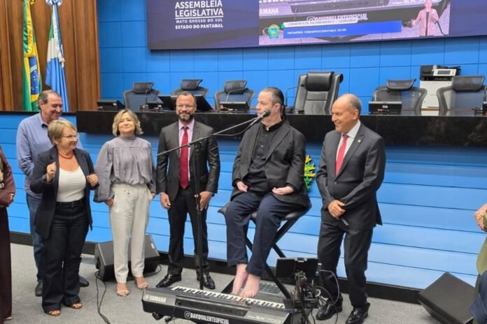 Group of five adults and two others stand in a blue legislative chamber; a man sits barefoot at a Yamaha keyboard, speaking into a microphone.