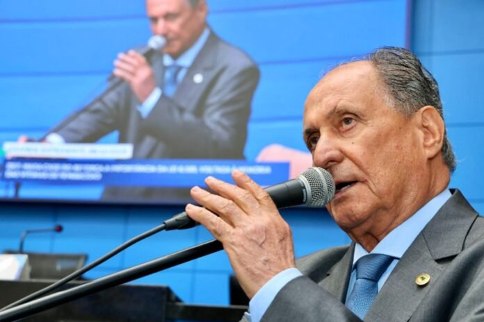 Older man in a gray suit and blue tie speaks into a handheld microphone on a stage, with a large blue screen showing his image behind him.