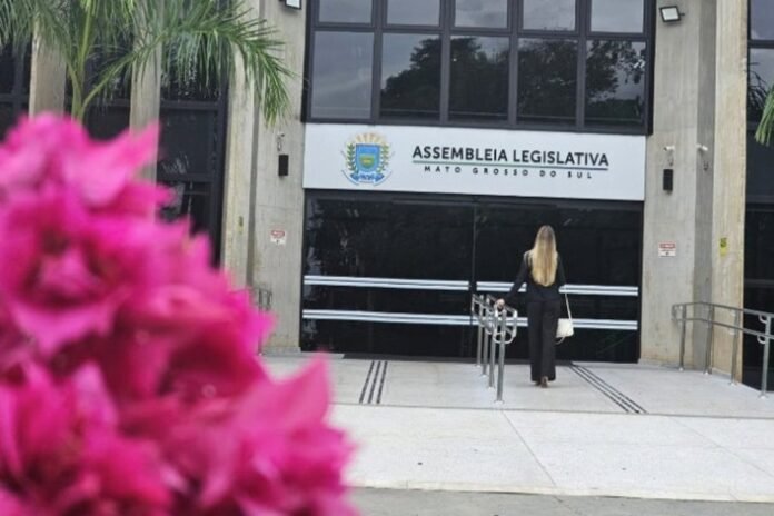 Woman in a black suit walking toward the entrance of the Assembleia Legislativa do Mato Grosso do Sul, with pink flowers in the foreground.