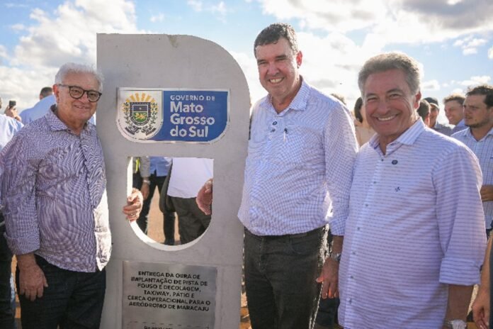Three men in light shirts stand beside a memorial plaque with the Mato Grosso do Sul government logo, smiling at a ceremony.