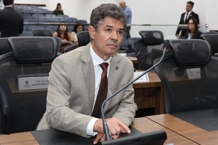 Man in a beige suit and red tie sits at a conference table with a microphone in front, in a formal meeting room.