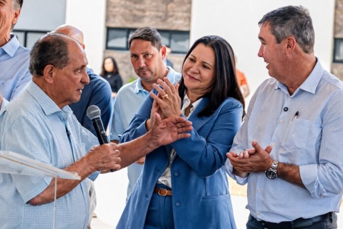 Man speaks into a microphone at an outdoor event while a woman in a blue suit and a man in a light shirt applaud nearby.