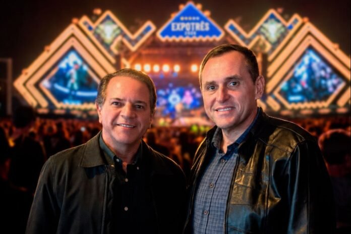 Two men pose for a photo at a crowded concert or expo venue, with a bright geometric stage backdrop in the background and a central EXPO sign behind them.
