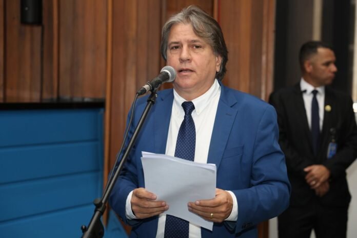 Man in a blue suit and white shirt speaks into a microphone, holding papers in a formal setting.