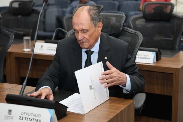 Older man in a dark suit at a conference table, turning through a white folder during a formal meeting.