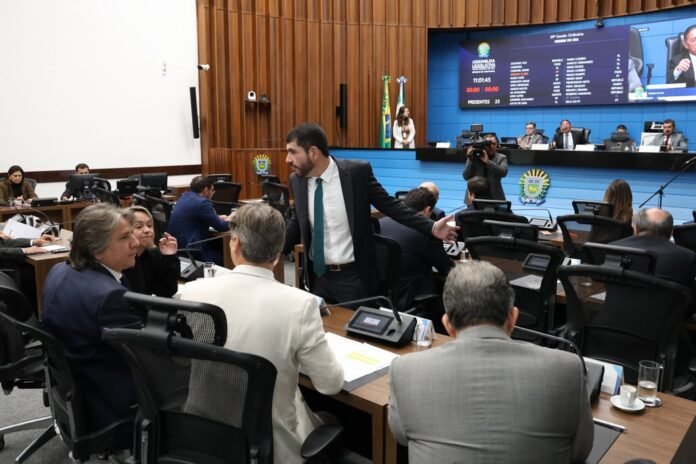 Politicians in a formal council chamber, a man in a suit stands and gestures while others listen at a long desk with microphones and documents.