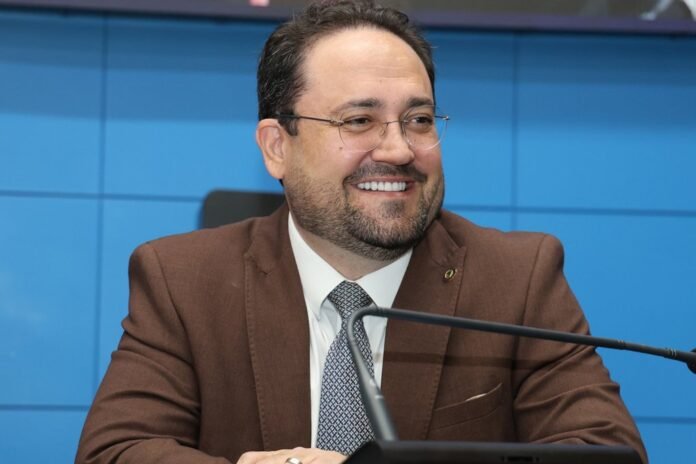 Smiling man in a brown suit and patterned tie sits at a panel with a microphone in front against a blue wall. (Note: concise alt text describing purpose.)